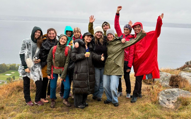 Eine Gruppe von zehn Personen steht auf einer grasbewachsenen Anhöhe mit Felsen, im Hintergrund ist ein großer See unter grauem, wolkenverhangenem Himmel zu sehen. Die Menschen tragen wetterfeste Kleidung wie Regenjacken, Ponchos und Mützen. Einige haben Rucksäcke, andere halten Getränke. Mehrere Personen heben die Arme fröhlich in die Luft, die Stimmung wirkt ausgelassen und gemeinschaftlich