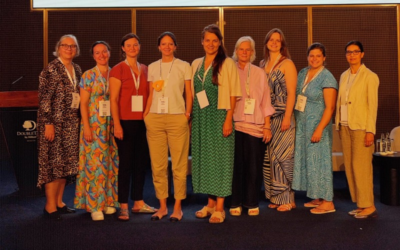 Neun Personen stehen gemeinsam auf einer Bühne bei einer Konferenz, alle tragen Namensschilder und sommerliche Kleidung