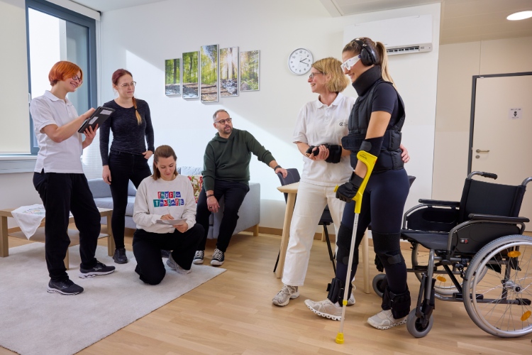 In einem hellen Therapieraum üben mehrere Personen gemeinsam das Gehen. Eine Patientin mit Augenbinde und Trainingsausrüstung wird dabei von einer Therapeutin begleitet. Zwei weitere Therapeutinnen sowie drei Personen beobachten und unterstützen die Übung. Neben der Patientin stehen ein Gehstock und ein Rollstuhl.