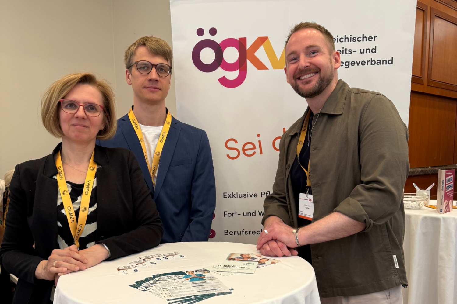 eine weibliche und zwei männliche Personen stehen an einem Stehtisch vor einem RollUp des ÖGKV