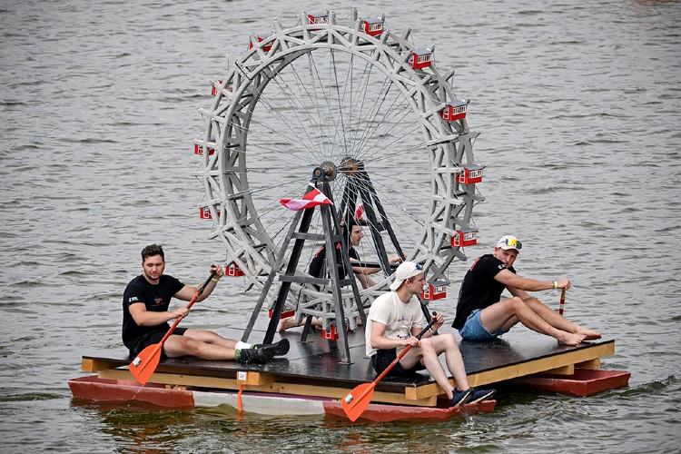 Vier Personen sitzen auf einem schwimmenden Floß und paddeln auf einem Gewässer. In der Mitte des Floßes befindet sich ein großes, detailreich nachgebildetes Wiener Riesenrad aus Beton. Die Konstruktion dient als ungewöhnliches Wasserfahrzeug und ist Teil eines Wettbewerbs. Im Hintergrund ist die Wasseroberfläche zu sehen.