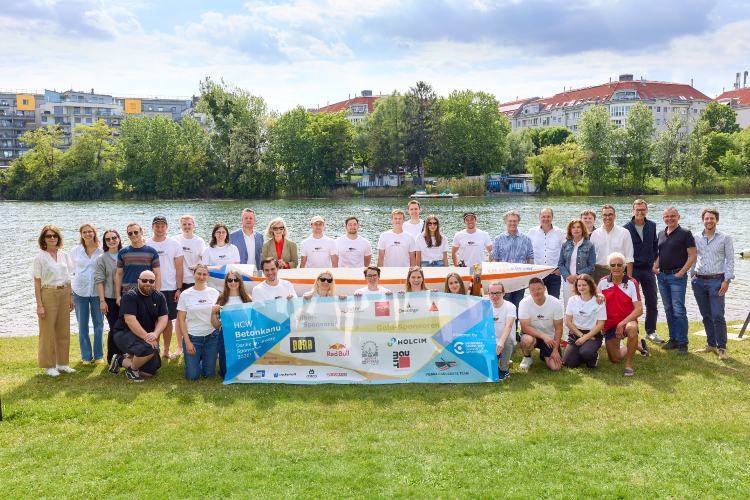 Gruppenfoto des Betonkanu-Teams der Hochschule Campus Wien an einem Flussufer. Rund 30 Personen stehen und knien gemeinsam auf einer Wiese hinter einem Banner mit Logos und der Aufschrift „HCW Betonkanu“. Im Hintergrund sind Wasser, Bäume und Gebäude zu sehen.